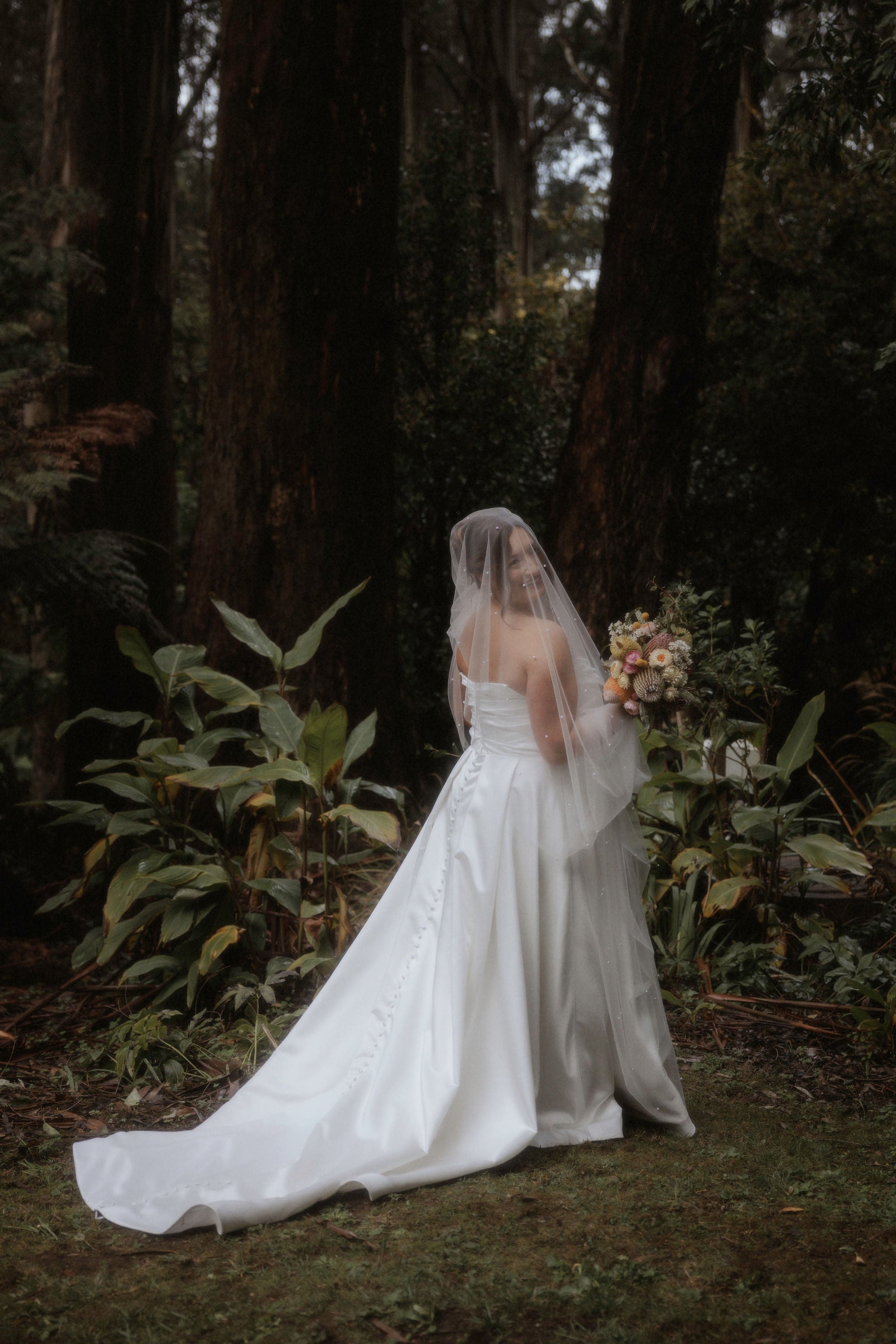 Bride in a white wedding dress standing in a forest setting with a veil and bouquet. Image by Who Shot The Photographer