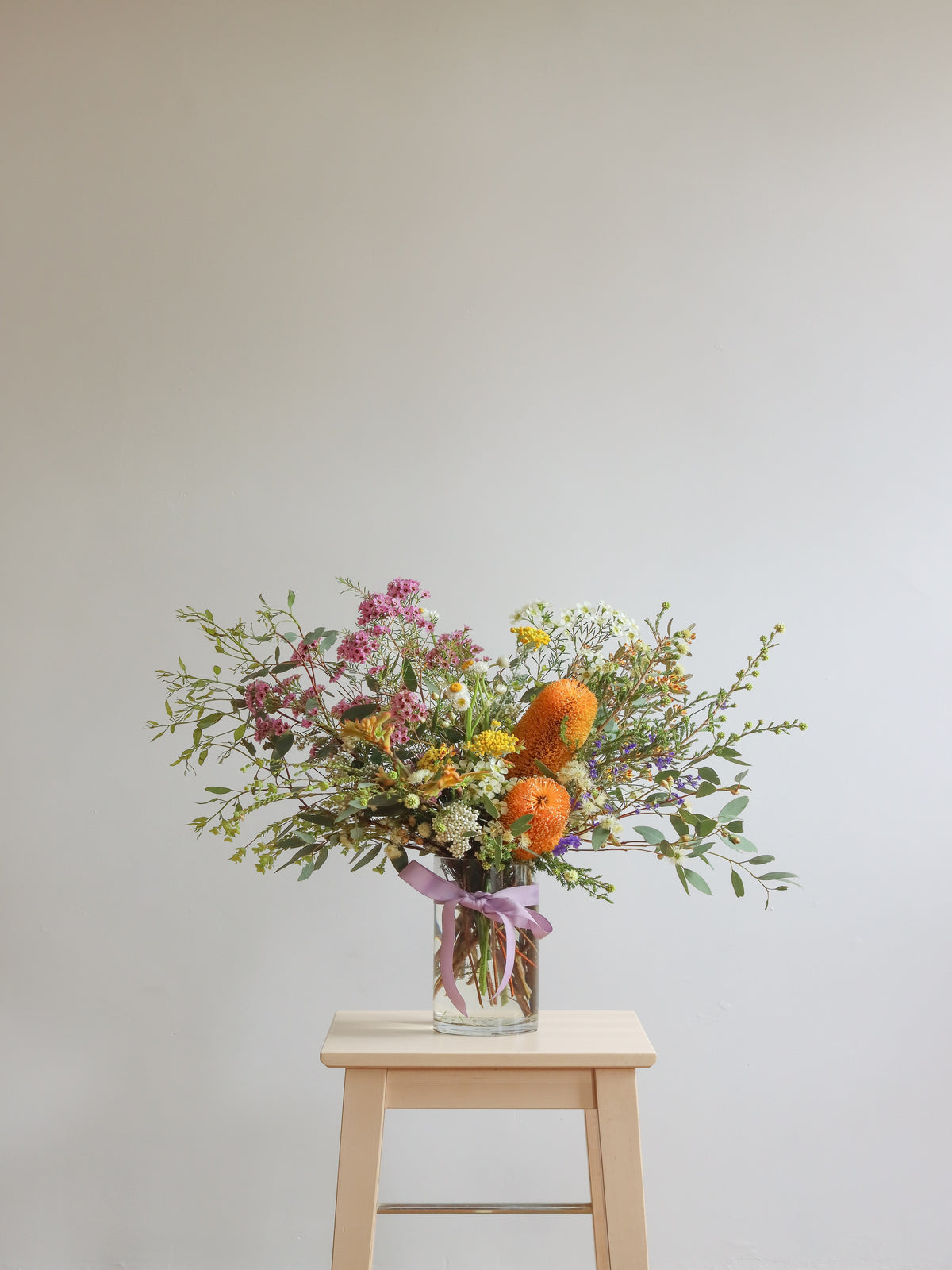 A wildflower bouquet in a clear glass jar vase, tied with a purple ribbon, displayed on a light wood stool against a neutral gray wall.