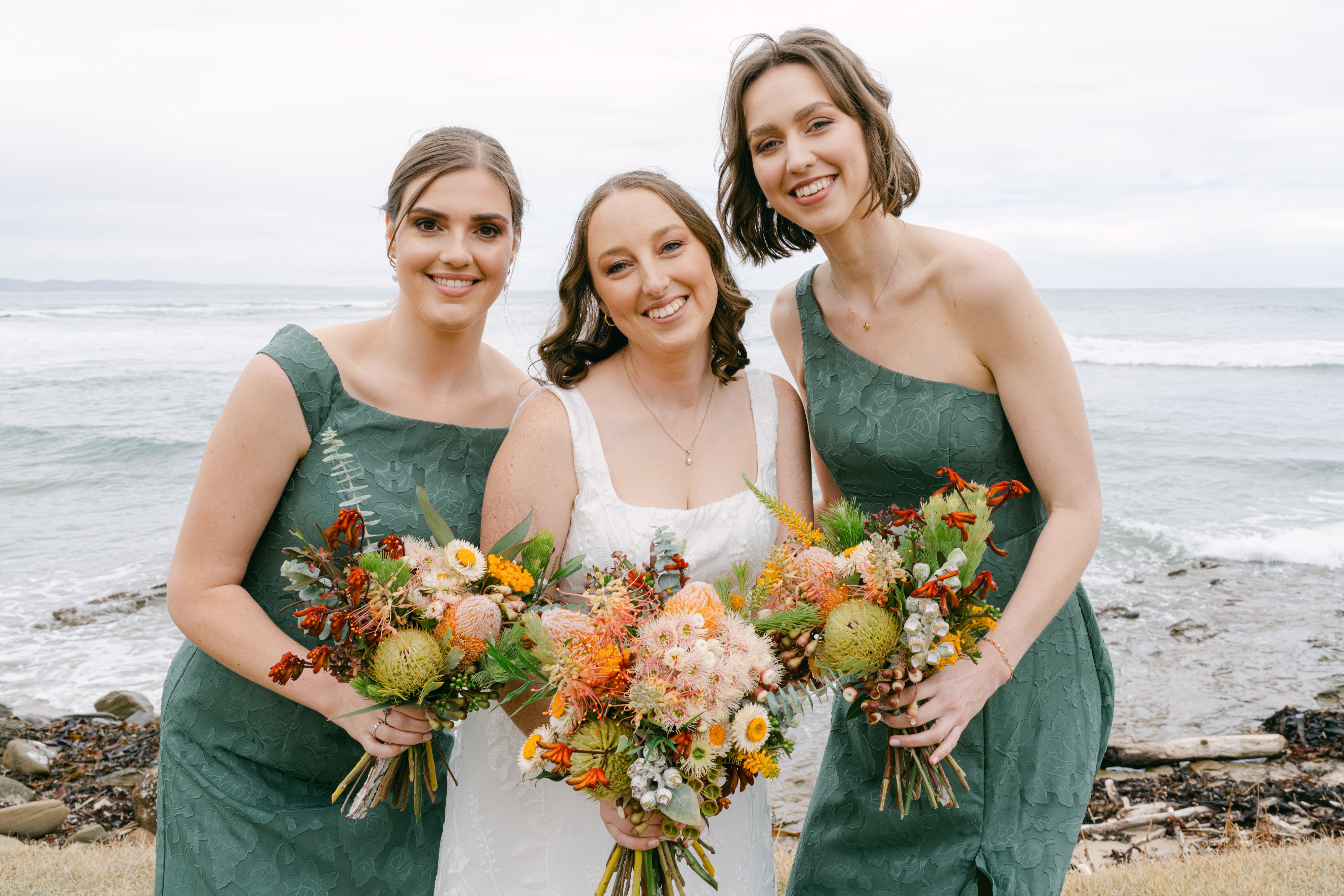 Bride in a white dress and two bridesmaids in green dresses holding native bouquets by the ocean.