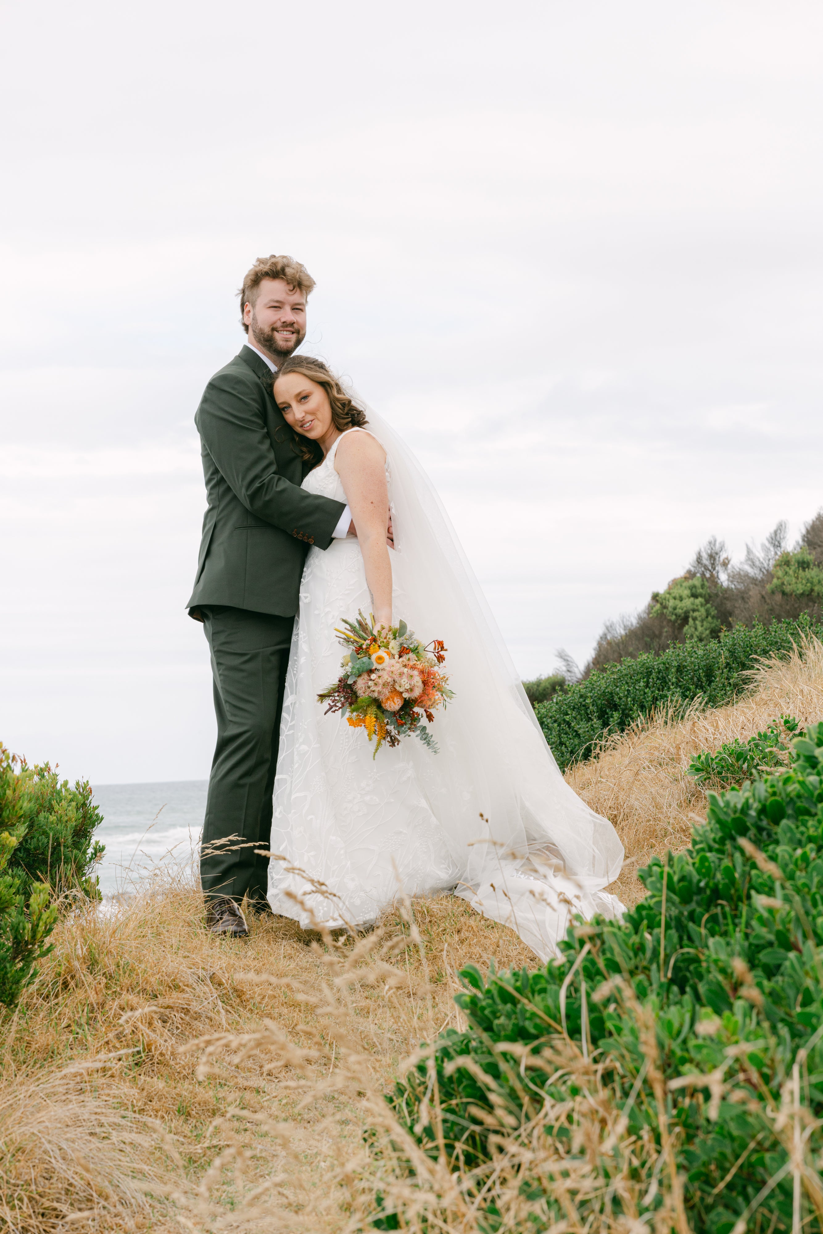 Couple in wedding attire standing on a beach with ocean view