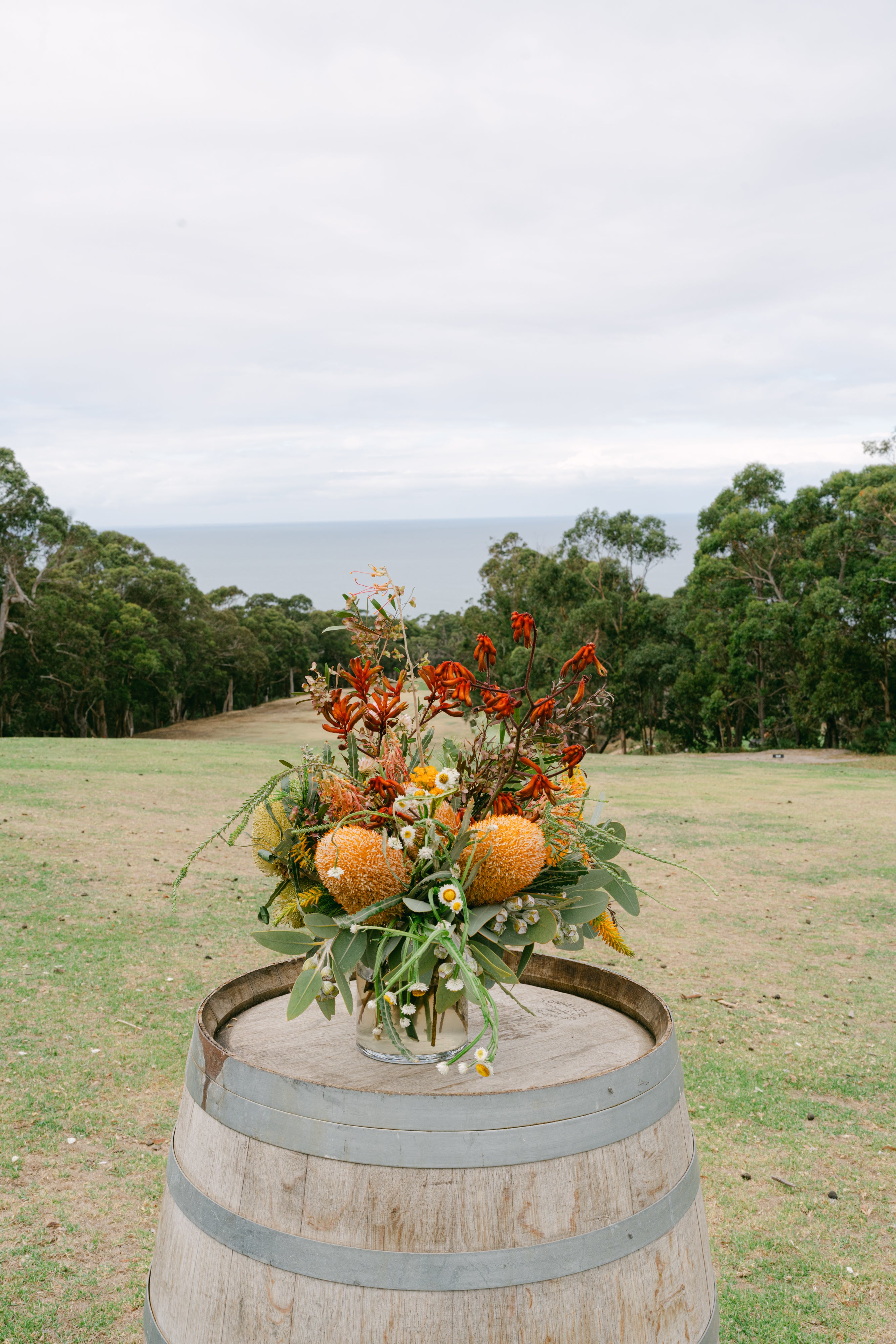 Decorative native floral arrangement on a wooden barrel with a scenic background of trees and sky.