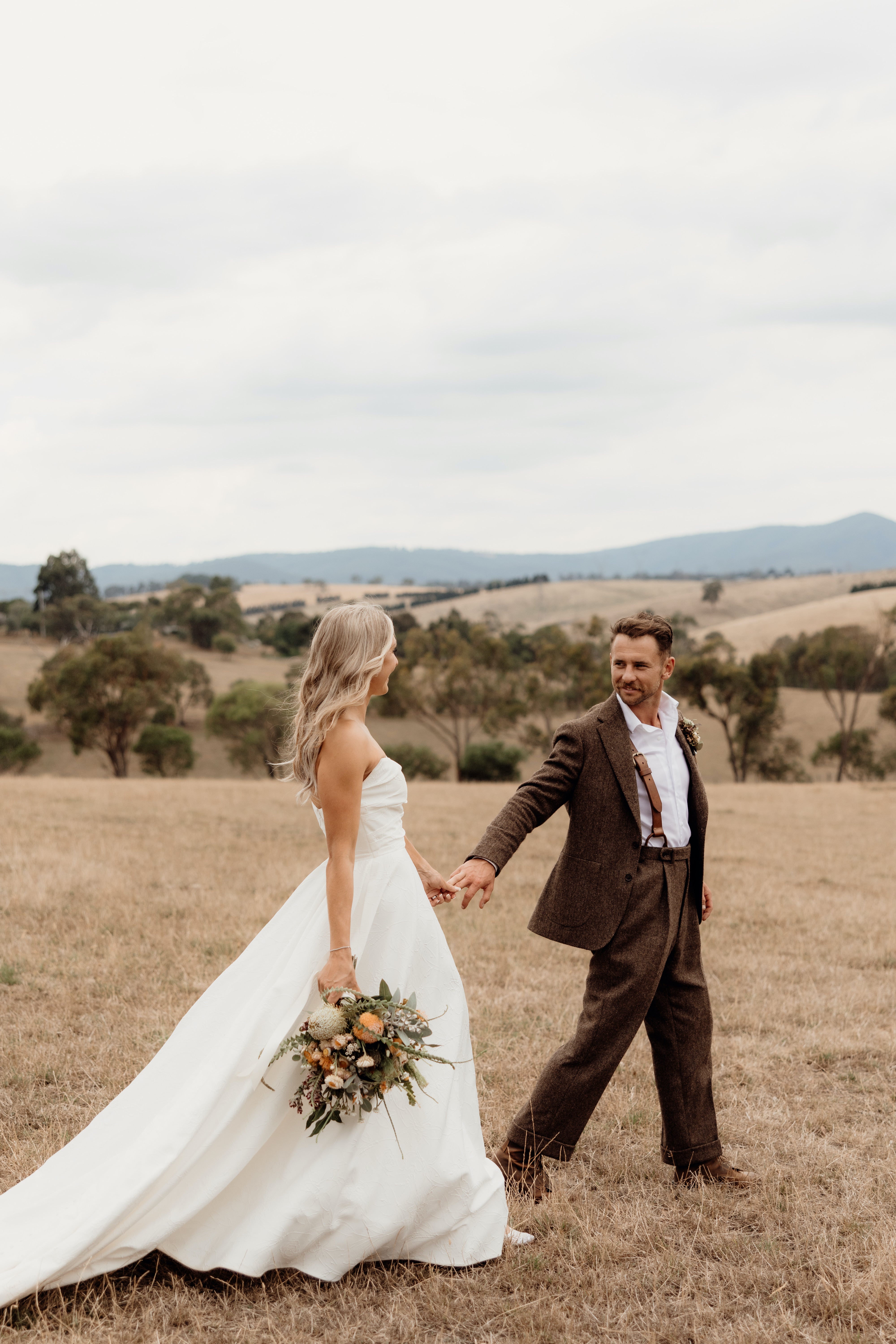 Couple in wedding attire holding hands in a field with a scenic background