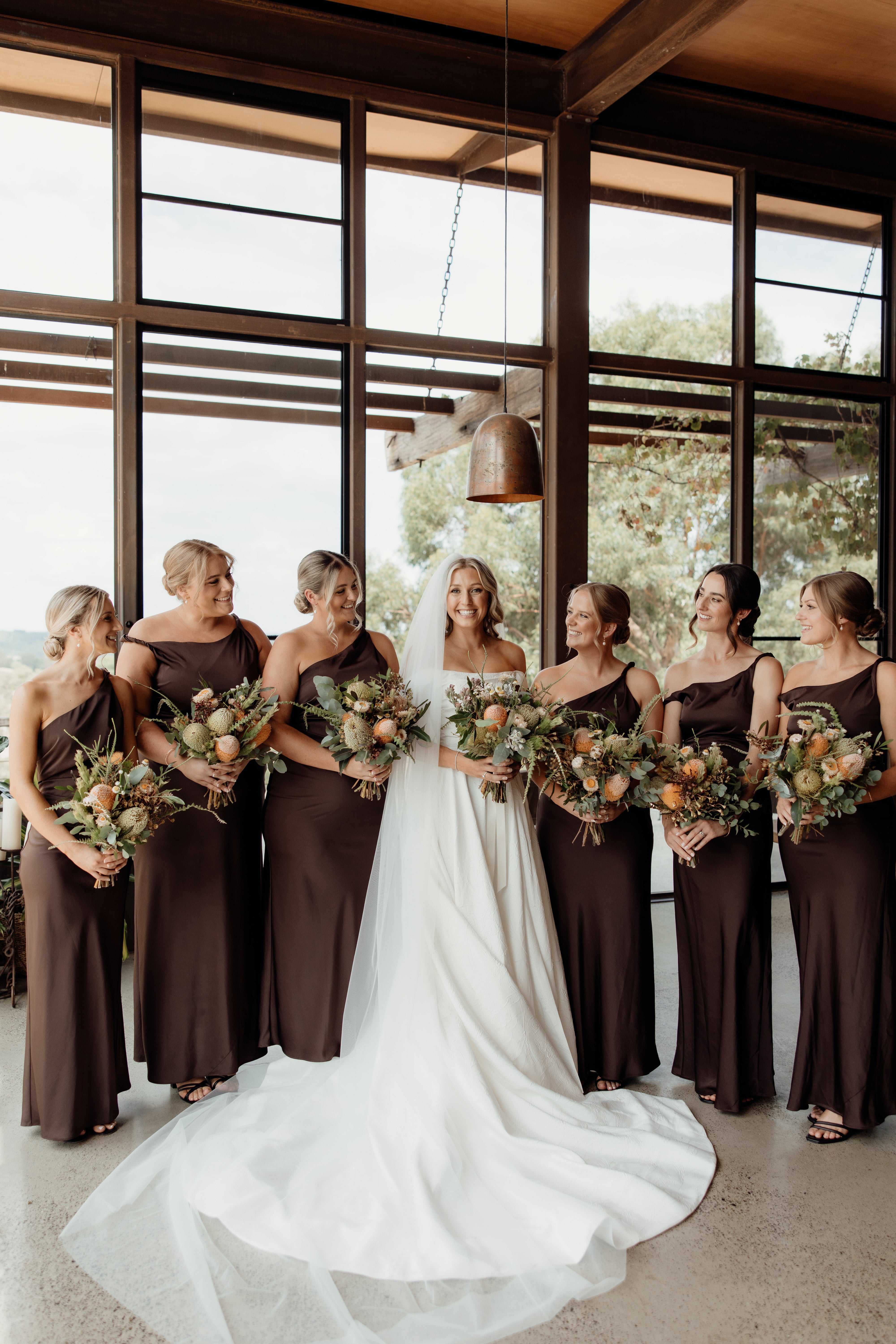 Bride with bridesmaids in brown dresses holding native floral bouquets indoors.