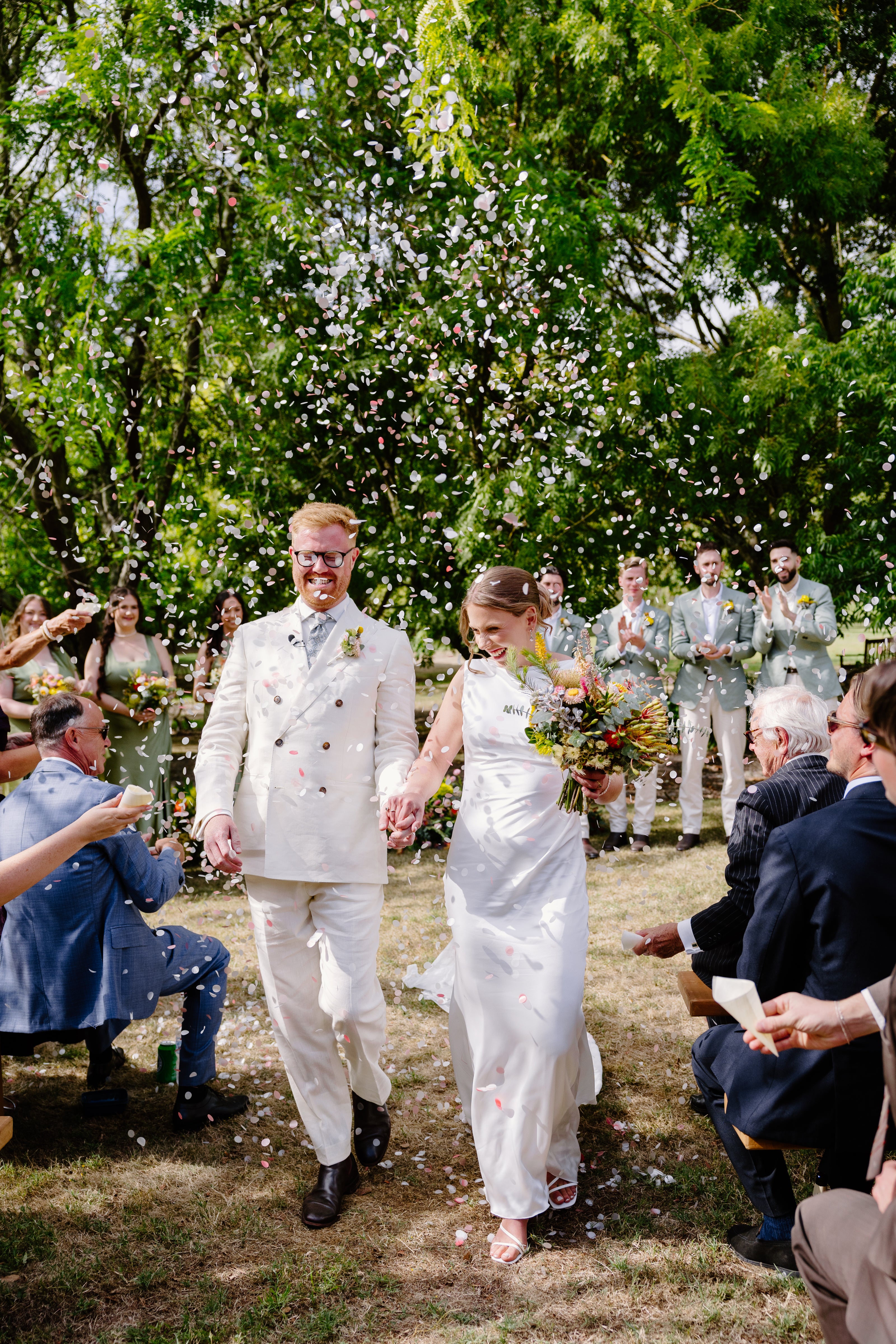 Wedding couple walking down an aisle with guests and confetti in an outdoor setting.