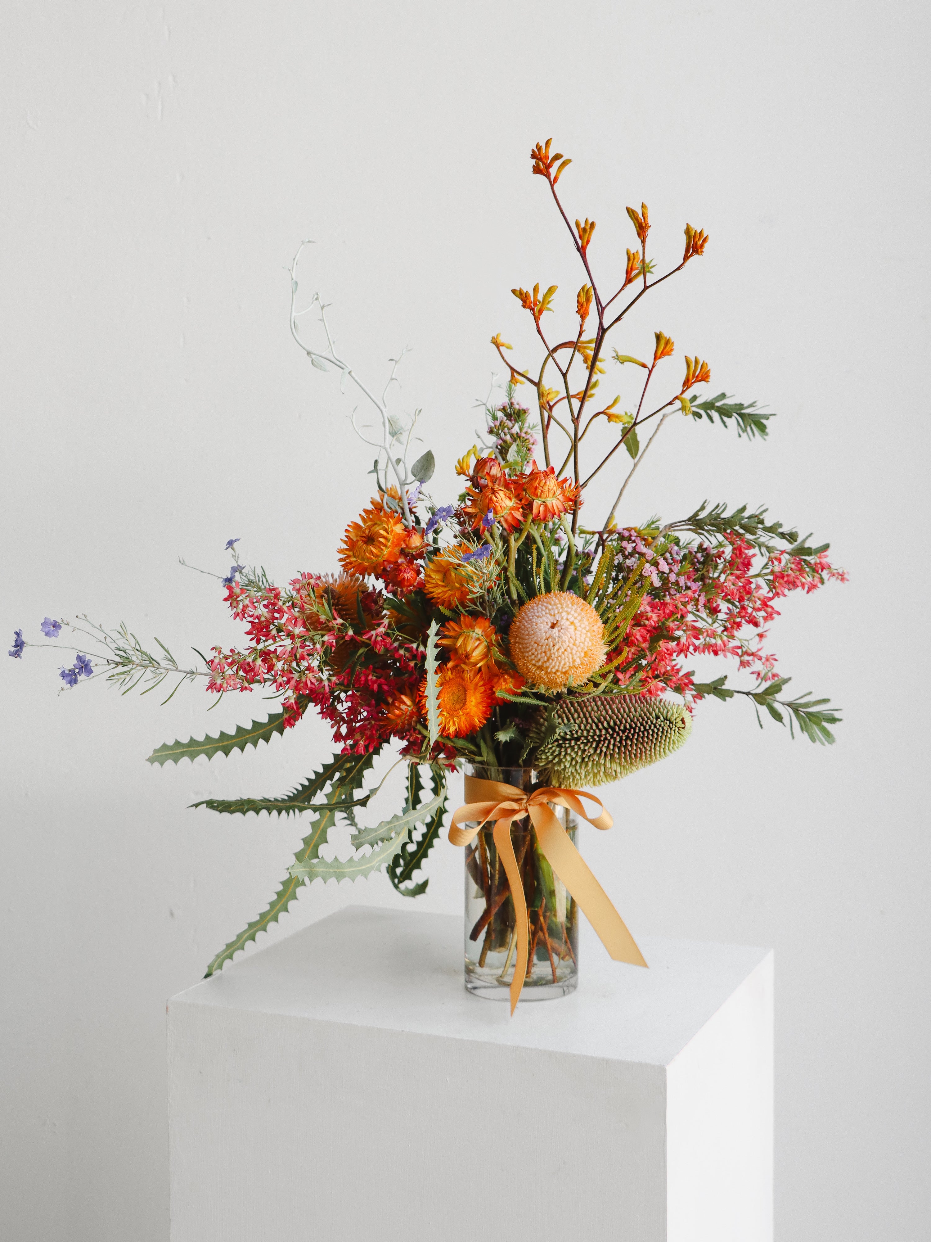 Bouquet of flowers with orange banksias in a clear vase on a white surface.