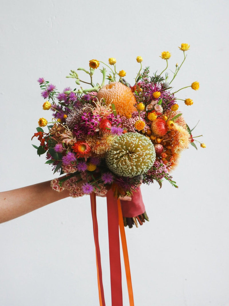 An example of a bush wedding bouquet, featuring banksia, orange flowering gum, yellow daisies, isopogon, purple waxflower, pink rice flower and melaleuca.